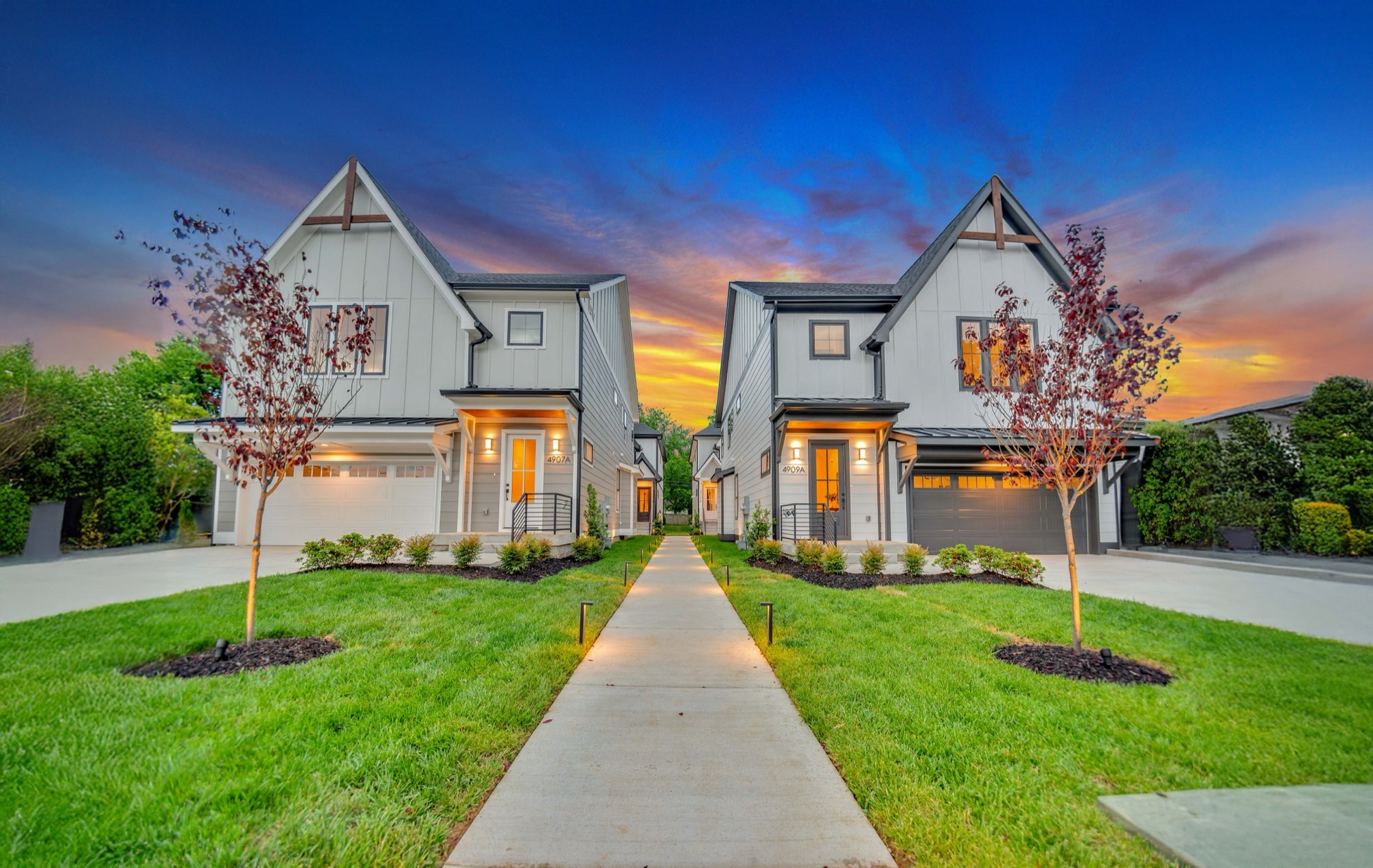 Two new farmhouse-style homes at sunset with a lit walkway between them